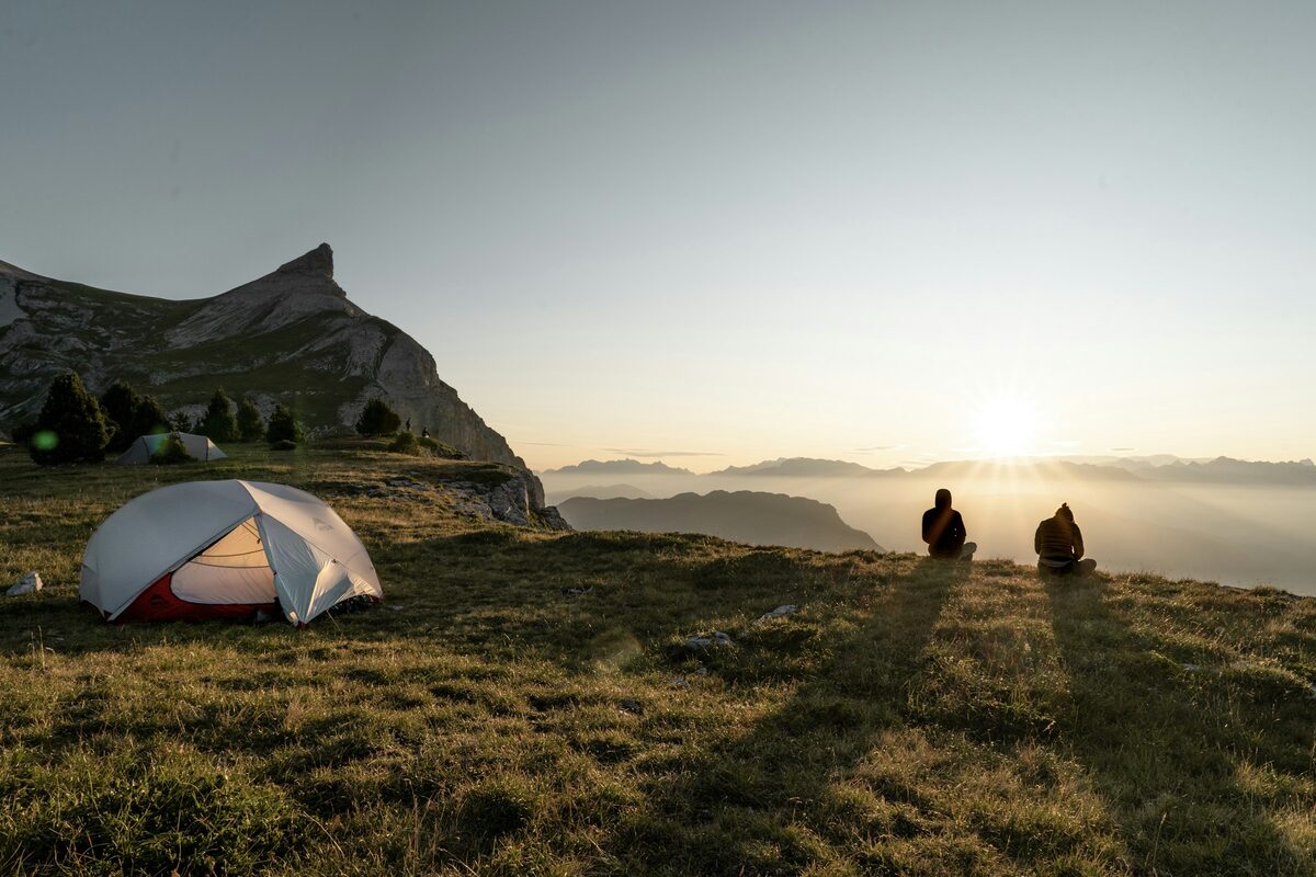 Tente dôme sur une crête herbeuse au lever du soleil, deux personnes assises face à la vallée