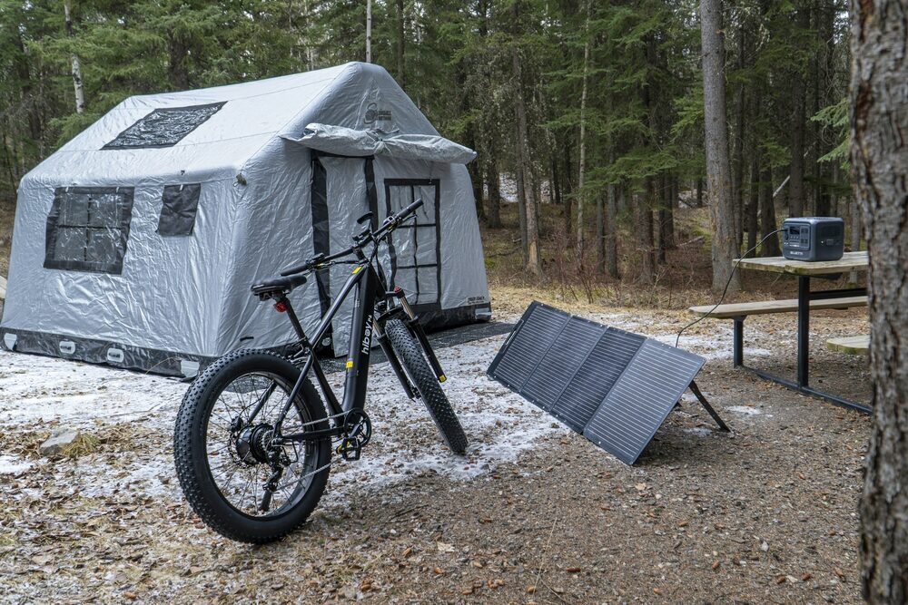 Tente gonflable grise, vélo et panneau solaire pliable posé au sol dans un camp en forêt
