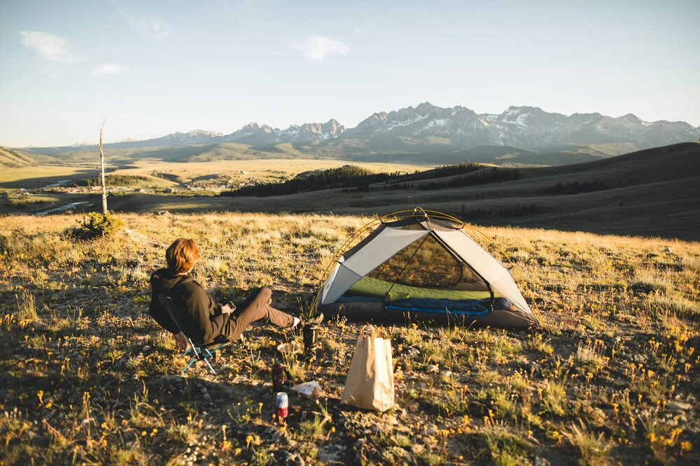 Personne assise dans l'herbe à côté d'une tente dôme, avec des montagnes au loin